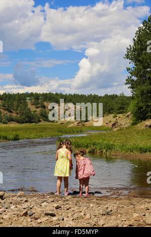 Zwei Schwestern stehen am Ufer eines Flusses auf der Suche nach unten ins Wasser mit großen bewaldeten Hügeln und blauen Himmel in der Ferne. Stockfoto