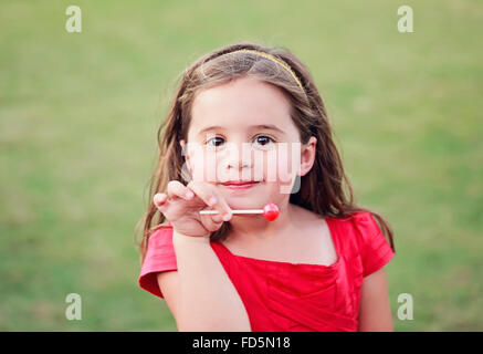 Kleines Mädchen in einem leuchtend roten Kleid einen leuchtenden roten Lutscher während lächelnd in die Kamera halten. Stockfoto