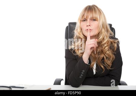 Schöne junge blonde Geschäftsfrau bitten um Geheimhaltung oder Stille halten den Finger an die Lippen, als sie an ihrem Schreibtisch sitzt, Stockfoto