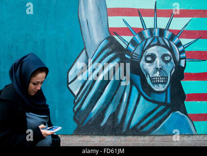 Frau mit Telefon vorbei an eine Anti-amerikanische Propaganda Slogan Darstellung Statue Freiheit Skelett an der Wand der Botschaft der Vereinigten Staaten, Central District, Teheran, Iran Stockfoto