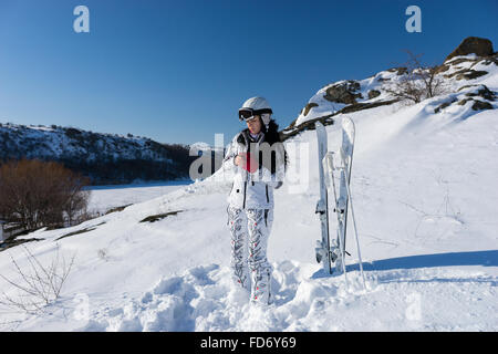 Voller Länge Candid junge Frau trägt weiße Skianzug und Helm anziehen rote Handschuhe und stehend nahe bei Ski und Stöcke auf Schnee bedeckt Berghang am hellen Tag mit Sonnenschein und blauem Himmel. Stockfoto