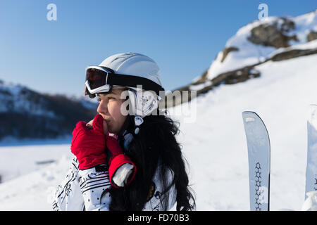 Kopf und Schultern Nahaufnahme junge Frau mit langen dunklen Haaren tragen Helm und Brille auf verschneiten Berg Blick in die Ferne und wärmende Hände mit Atem an sonnigen Tag. Stockfoto