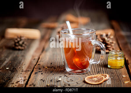 Ein Glas heißer schwarzer Tee, Tannenzapfen, ein wenig Honigtopf und Orangenscheiben auf einem Holztisch Stockfoto