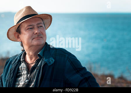 Erwachsener Mann mit einem Schnurrbart und Bart in einen Strohhut gegen das Meer lächelnd. Close-up Portrait eines älteren Mannes. Sonnigen Tag Stockfoto