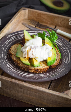 Pochiertes Ei und Avocado auf geröstetem Vollkornbrot mit Samen auf einem Teller in Vintage Holztablett. Garniert mit Frühlingszwiebeln Stockfoto