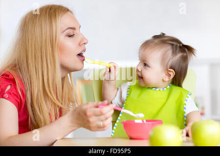 Kleine Mädchen und Mutter mit Baby-Nahrung füttern einander, sitzt am Tisch im Kindergarten Stockfoto