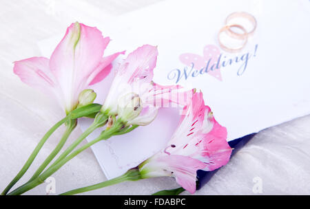Schöne rosa Lilien ruhen auf einer Hochzeitseinladung mit Trauringe unscharf im Hintergrund. Stockfoto