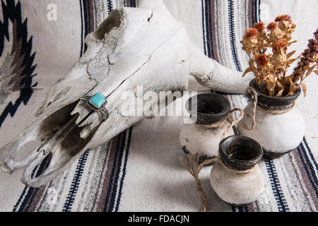 Verwitterter Kuhschädel, türkisfarbenes und silbernes Manschettenarmband, schwarz-weiß bemalte mexikanische Keramik mit getrockneten Blumen auf einer mexikanischen gestreiften Decke. Stockfoto