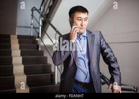 Geschäftsmann mit Handy in der Hand macht rufen während auf Treppen an Bürogebäude in Pause, Anzug. Business Portrait eines männlichen Mitte-asiatischen Aussehens. Stockfoto
