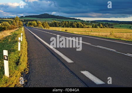 Asphaltierte Straße durch die Felder bis zum Horizont, bewaldeten Berge am Horizont, stürmischen Wolken Stockfoto