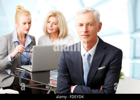 Porträt von senior Geschäftsmann im Büro während seiner Kollegen arbeiten im Hintergrund am Laptop sitzt. Stockfoto