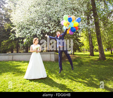 Bräutigam auf die Ballons fliegen, aber die Braut hält seine Hand. Lustige Hochzeit. Bräutigam springt und fliegt auf Ballons. Junger Mann von seiner Frau fliegen. Lustige Hochzeitskonzept. Happy Valentines day Stockfoto