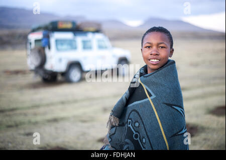 Porträt eines jungen Mannes, eingewickelt in eine Decke mit Bergen und einem Landrover in Sani Pass, Lesotho, Afrika im Hintergrund. Stockfoto