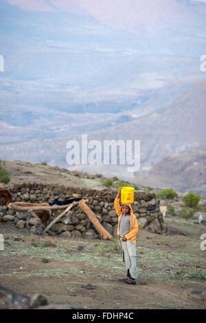 Kleiner Junge mit einem Eimer auf dem Kopf vor einer Bergkette in seinem Dorf in Lesotho, Afrika Stockfoto