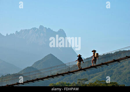 Zwei Bauern, die zu Fuß über eine Hängebrücke in Kota Belud, Sabah mit seitlichem Blick auf den Mount Kinabalu im Hintergrund. Stockfoto