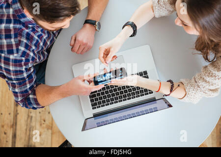 Draufsicht der junge Mann und Frau mit Laptop und zeigt Bildschirm des Smartphones am runden Tisch sitzen Stockfoto