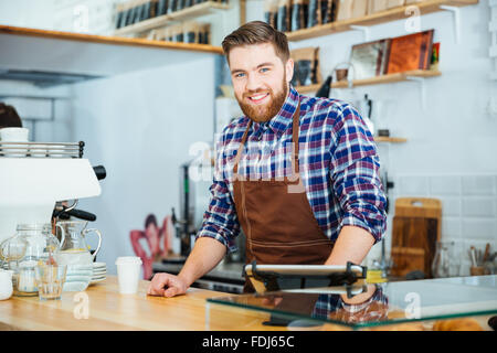 Fröhliche hübscher Junge Barista mit Bart in braun Schürze im Coffee-shop Stockfoto