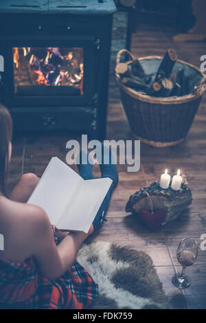 Frau sitzt vor einem hölzernen brennenden Ofen mit einem Buch Stockfoto