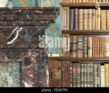 Detail einer Ecke am Kamin, ein Teil der Bücherregale und der viktorianischen Cordelova geprägte Tapete in der Bibliothek im Dunster Castle, Somerset. Stockfoto