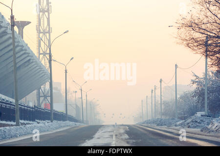 Der Hintergrund, auf einer leeren Straße in der Stadt am kalten Wintertag mit Schnee in der Nähe von Stadion, Luftperspektive bedeckt. Lichtmasten an Rändern der Straße. Stockfoto