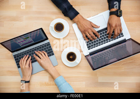 Draufsicht der Hände von afrikanischen Mann und kaukasischen Frau Tippen auf zwei Laptops und Kaffeetrinken auf Holztisch Stockfoto