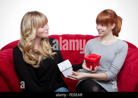 schöne junge blond und rot behaarte Mädchen geben Geschenkbox auf roten Sofa vor weißem Hintergrund Stockfoto