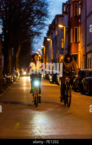 Radfahrer fahren, in der Dämmerung, in einer Stadtstraße, mit und ohne Beleuchtung und Sicherheitskleidung, Fahrrad Sicherheit, Sichtbarkeit bei Nacht Stockfoto