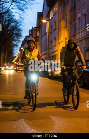 Radfahrer fahren, in der Dämmerung, in einer Stadtstraße, mit und ohne Beleuchtung und Sicherheitskleidung, Fahrrad Sicherheit, Sichtbarkeit bei Nacht Stockfoto