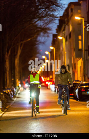 Radfahrer fahren, in der Dämmerung, in einer Stadtstraße, mit und ohne Beleuchtung und Sicherheitskleidung, Fahrrad Sicherheit, Sichtbarkeit bei Nacht Stockfoto