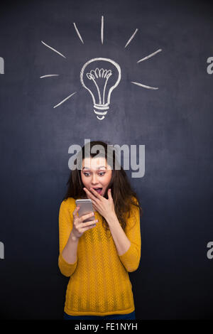 Glückliche junge Frau erstaunt mit geöffneten Mund mit Handy über Tafel Hintergrund Stockfoto