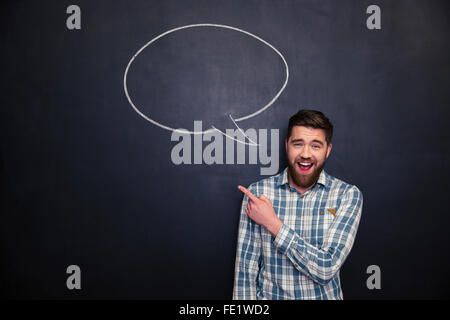 Fröhliche Jüngling bärtigen zeigenden Weg stehend über Blackboard Hintergrund mit leere Sprechblase Stockfoto