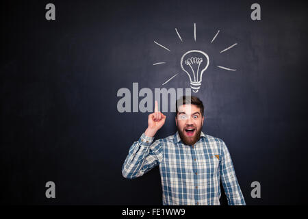 Aufgeregt bärtiger junger Mann im karierten Hemd nach oben über Blackboard Hintergrund mit gezeichneten Glühbirne Stockfoto