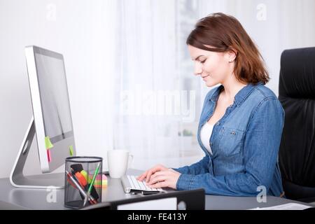 Lächelnde Geschäftsfrau, die Eingabe von Daten auf ihrem Desktop-Computer sitzen Tippen auf der Tastatur, Seitenansicht Stockfoto