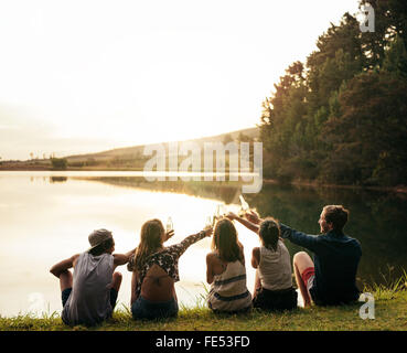 Gruppe junger Leute sitzen in einer Reihe an einem See. Junge Freunde, toasten und mit Bier am See zu feiern. Stockfoto