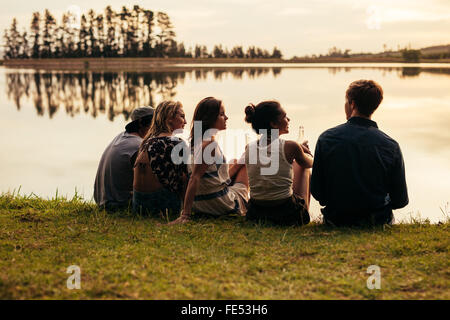 Hintere Ansicht Porträt Gruppe von jungen Freunden entspannende an einem See. Junge Menschen sitzen zusammen an einem See. Stockfoto