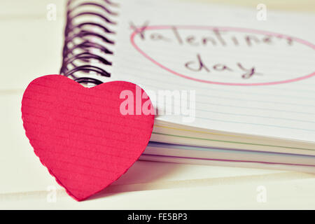 Nahaufnahme von einem roten Herz und ein Notebook mit der Text-Valentinstag handgeschrieben, auf einer weißen Fläche, mit einem Filtereffekt Stockfoto