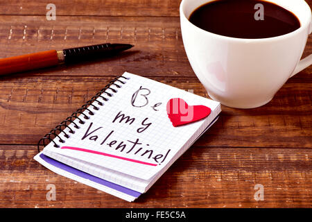 ein Notebook mit dem Text werden mein Valentinsgruß handschriftlich in it, ein rotes Herz, einen Stift und eine Tasse Kaffee auf einem rustikalen Holztisch Stockfoto