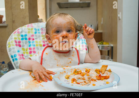 Ein kleines Mädchen (Alter 15 Monate) isst eine Mahlzeit aus Mais und Tomatensauce in gewissem Sinne chaotisch und spielt mit ihr Essen auf einem hohen Stuhl Stockfoto