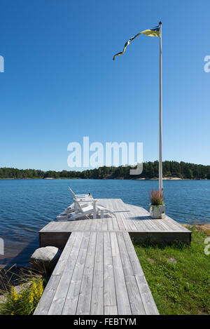 Holzsteg und Belag mit zwei weißen Liegestühlen, Fahnenmast und schwedische farbige Flagge auf der Insel an der Ostsee, Schweden Stockfoto