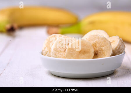 In Scheiben geschnittenen Bananen in einer weißen Schüssel auf einen weißen Tisch vor zwei Bananen mit gelben Schalen. Stockfoto