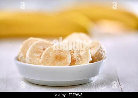 Frisch geschnittene Bananenscheiben in einer weißen Schüssel auf einem Holztisch vor gelben Bananenschalen Stockfoto