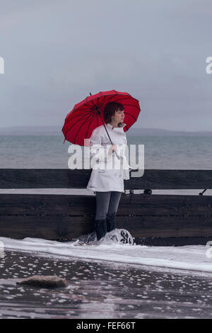 eine Frau in einem weißen Mantel mit einem roten Regenschirm am Meer im winter Stockfoto