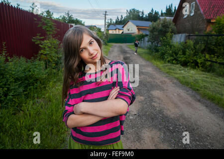 Lächeln Teenager-Mädchen in einem roten gestreiftes Hemd steht auf der Straße zwischen den Häusern, verschränkte ihre Arme über der Brust. Stockfoto