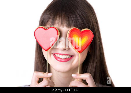 Kopfschuss-Portrait von junge schöne verspielte Frau halten, dass zwei herzförmige rote Cookies, herumalbern, mit Keksen als Augen Stockfoto