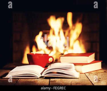 Rote Tasse Kaffee oder Tee und alte Bücher auf Holztisch in der Nähe von Kamin. Winter und Weihnachten Ferienkonzept. Foto mit Retro-f Stockfoto