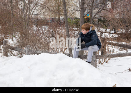 Kleiner Junge sitzt auf einem hölzernen Zaun hält einen Schneeball Stockfoto