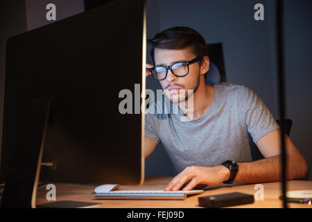 Nachdenklicher junger Mann in Gläsern denken und arbeiten mit Computer im dunklen Raum Stockfoto