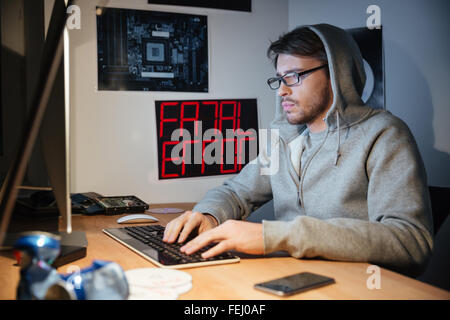 Hübscher junger Mann in Sweatshirt mit Kapuze sitzen und schreiben auf der Computertastatur zu Hause Stockfoto