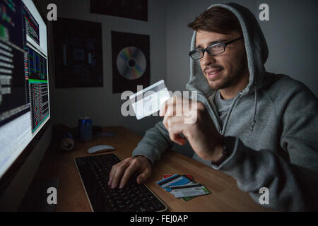Lächelnde junge Kautschuk im Sweatshirt mit Kapuze, Einkaufen im Internet und bezahlen mit gestohlenen Kreditkarte Stockfoto