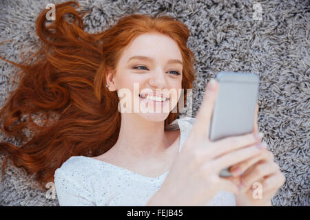 Lächelnde rothaarige Frau liegend auf dem Teppich und mittels Smartphone zu Hause Stockfoto